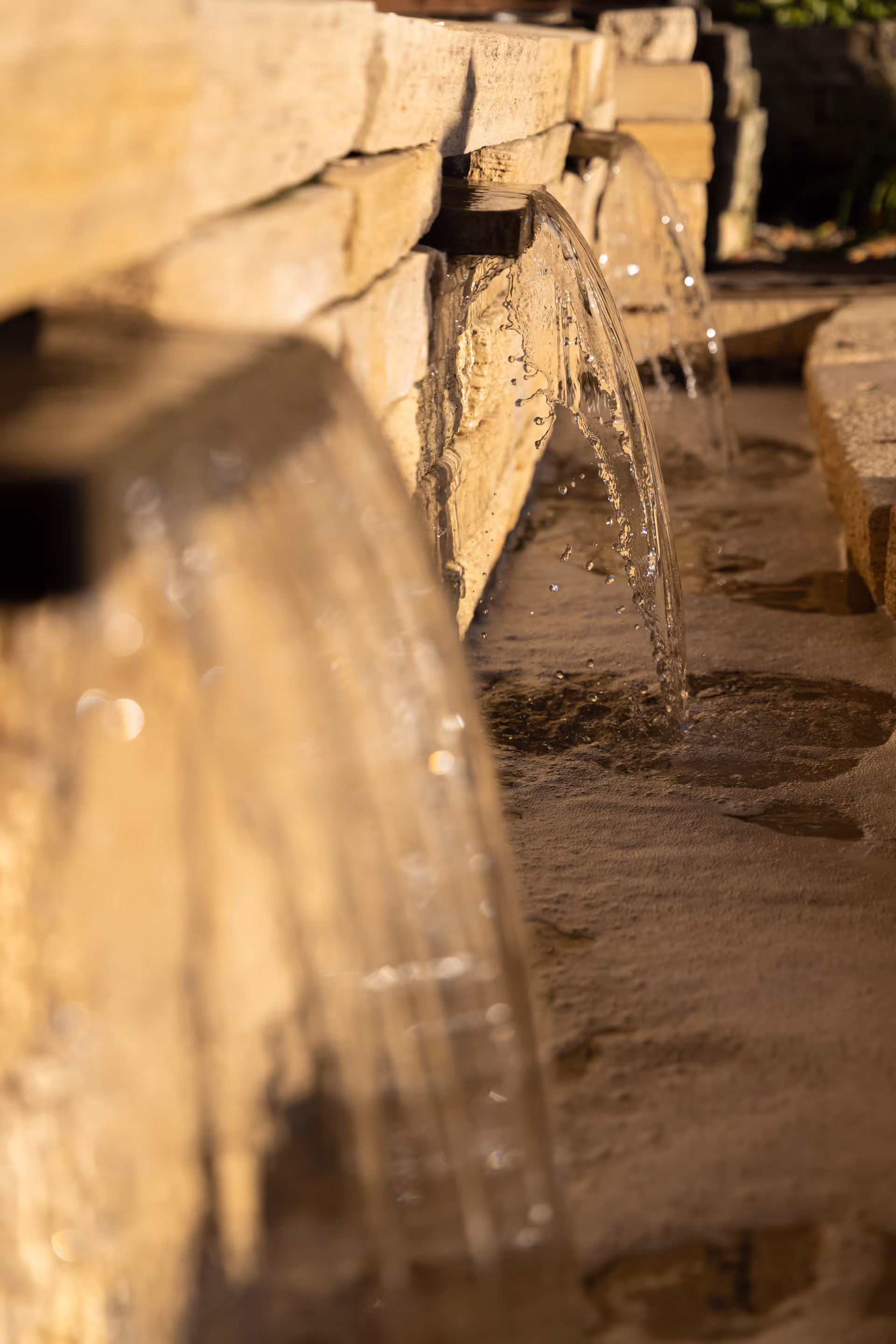 Close-up view of water flowing from spouts built into a stone wall into a shallow basin, with sunlight illuminating the water and stone surface.