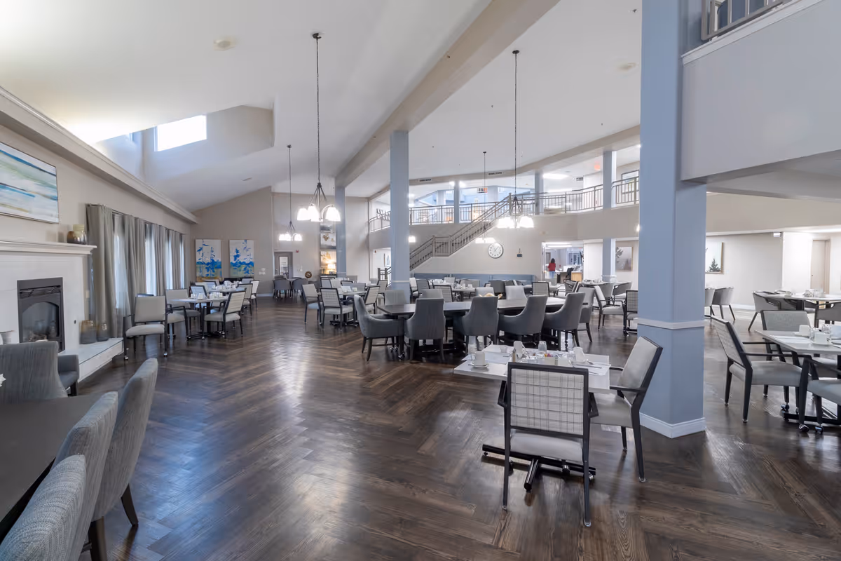 Spacious dining room with multiple tables and chairs arranged neatly. The room features high ceilings with hanging light fixtures, large windows with curtains, a fireplace on the left wall, and a staircase leading to an upper level. The floor is dark wood with a herringbone pattern, and the walls are painted in light neutral tones. The tables are set with white cups and plates.