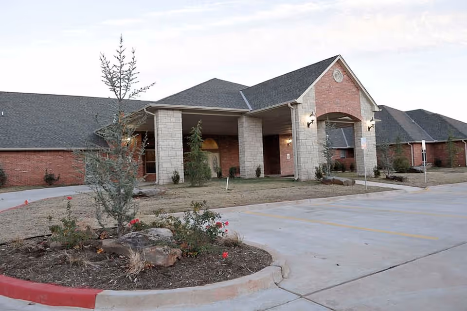 Exterior view of Tealridge Assisted Living and Memory Care facility showing a single-story building with a covered entrance supported by stone columns, a landscaped area with small trees and bushes, and a concrete driveway and parking area in front.