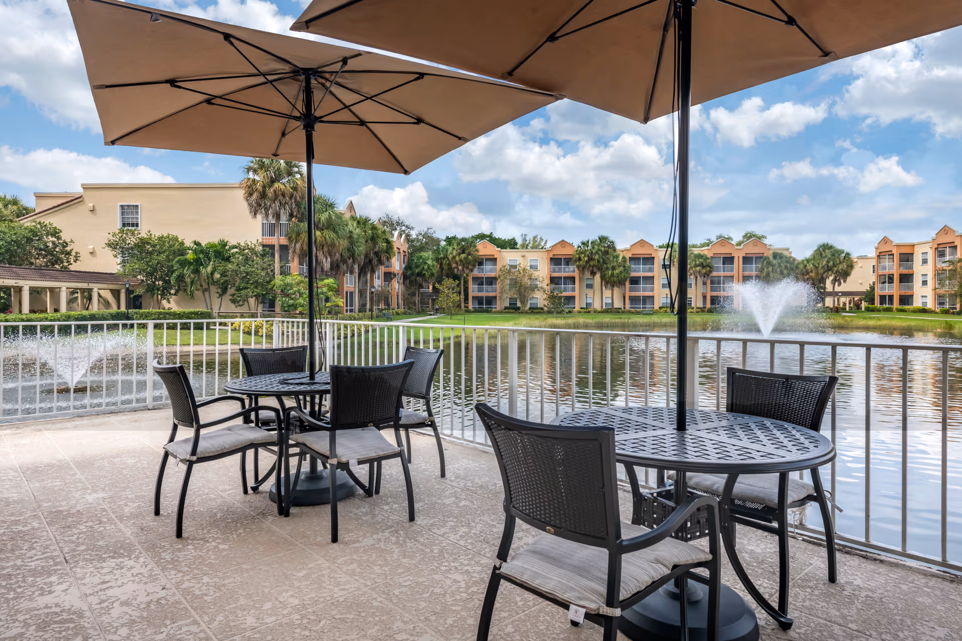 Outdoor patio with tables, chairs and umbrellas overlooking a pond with fountains and apartment-style buildings in the background.
