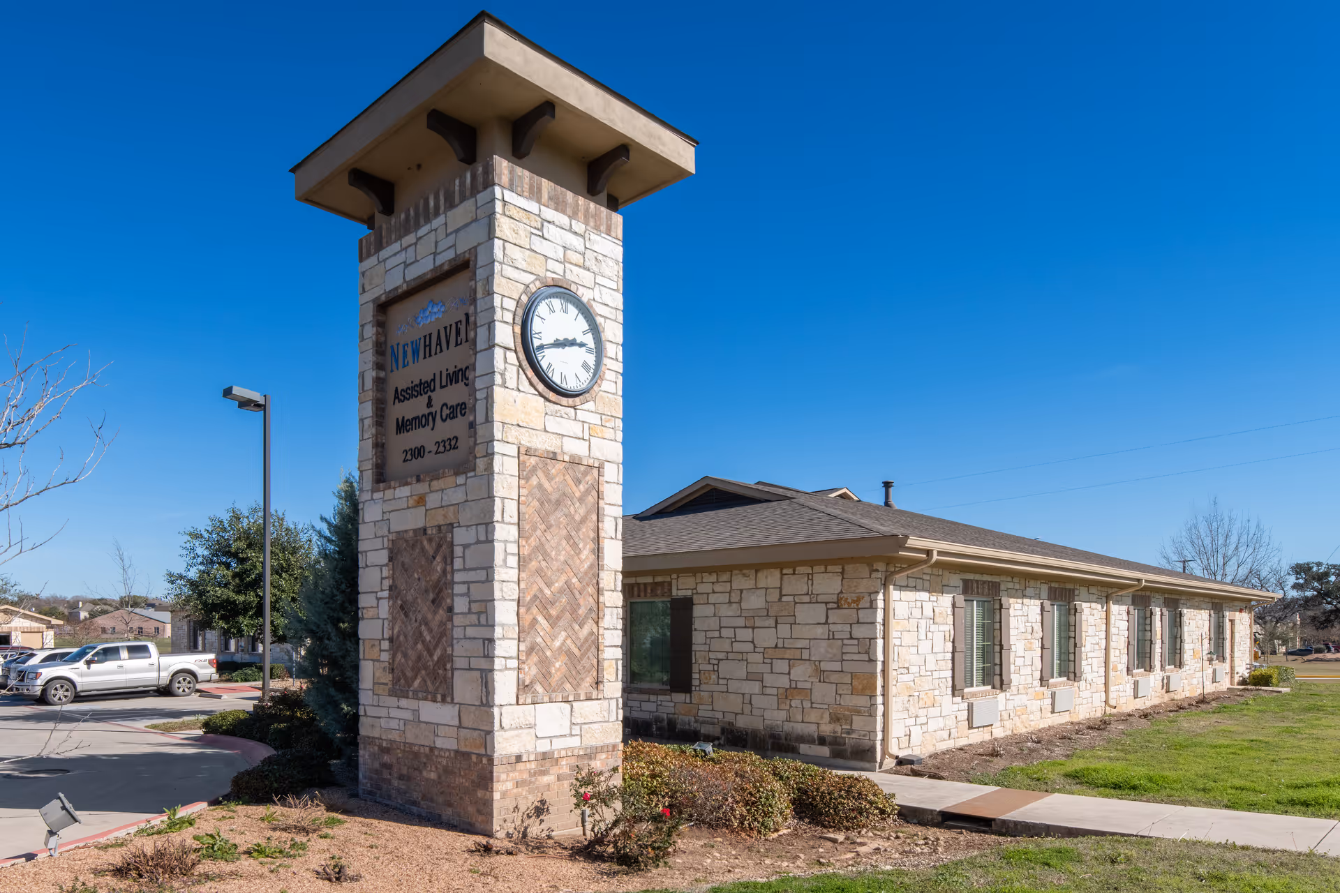 Exterior view of New Haven Assisted Living and Memory Care of Schertz building with a tall stone clock tower displaying the facility name and address. The building is made of light-colored stone with multiple windows and is surrounded by a grassy area and a parking lot with vehicles.