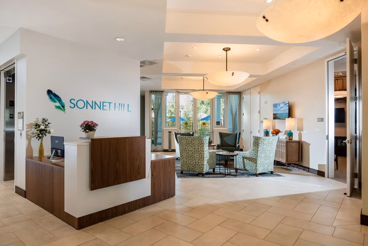 Reception area of Sonnet Hill Senior Living featuring a wooden front desk with a computer and flower vases. Behind the desk is a wall with the Sonnet Hill logo and a decorative feather. The seating area includes patterned armchairs around a coffee table, with large windows and curtains letting in natural light. The space is well-lit with ceiling lights and has a tiled floor.