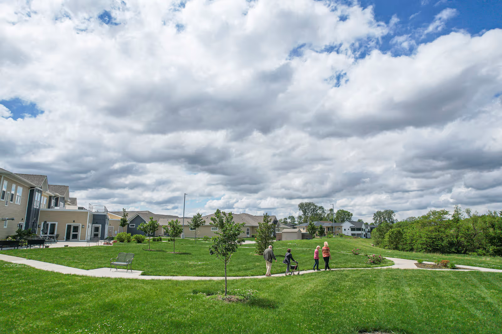 Green lawn and walking paths at a senior living community with several residents walking near low-rise buildings under a cloudy sky.
