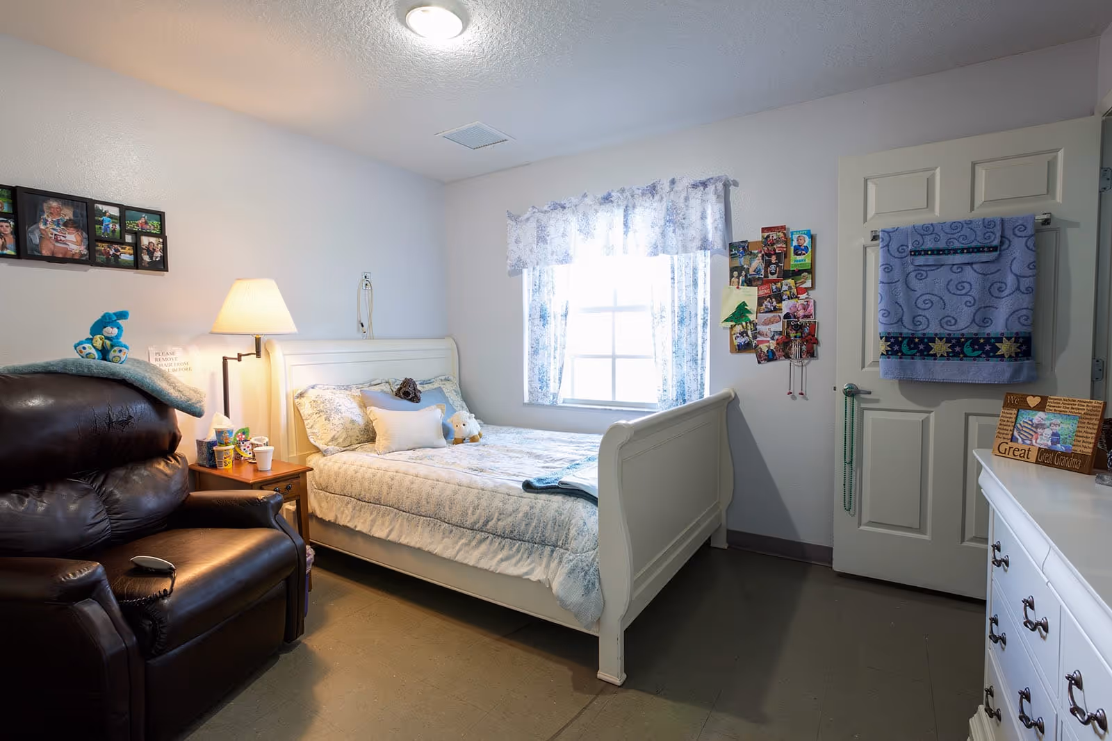 A cozy bedroom in an assisted living facility featuring a white wooden bed with floral bedding and stuffed animals, a brown leather recliner chair with a blue blanket and stuffed toy on it, a wooden nightstand with a lamp and personal items, a window with floral curtains, a door with a blue towel hanging on it, and a white dresser with a framed photo and decorative items.