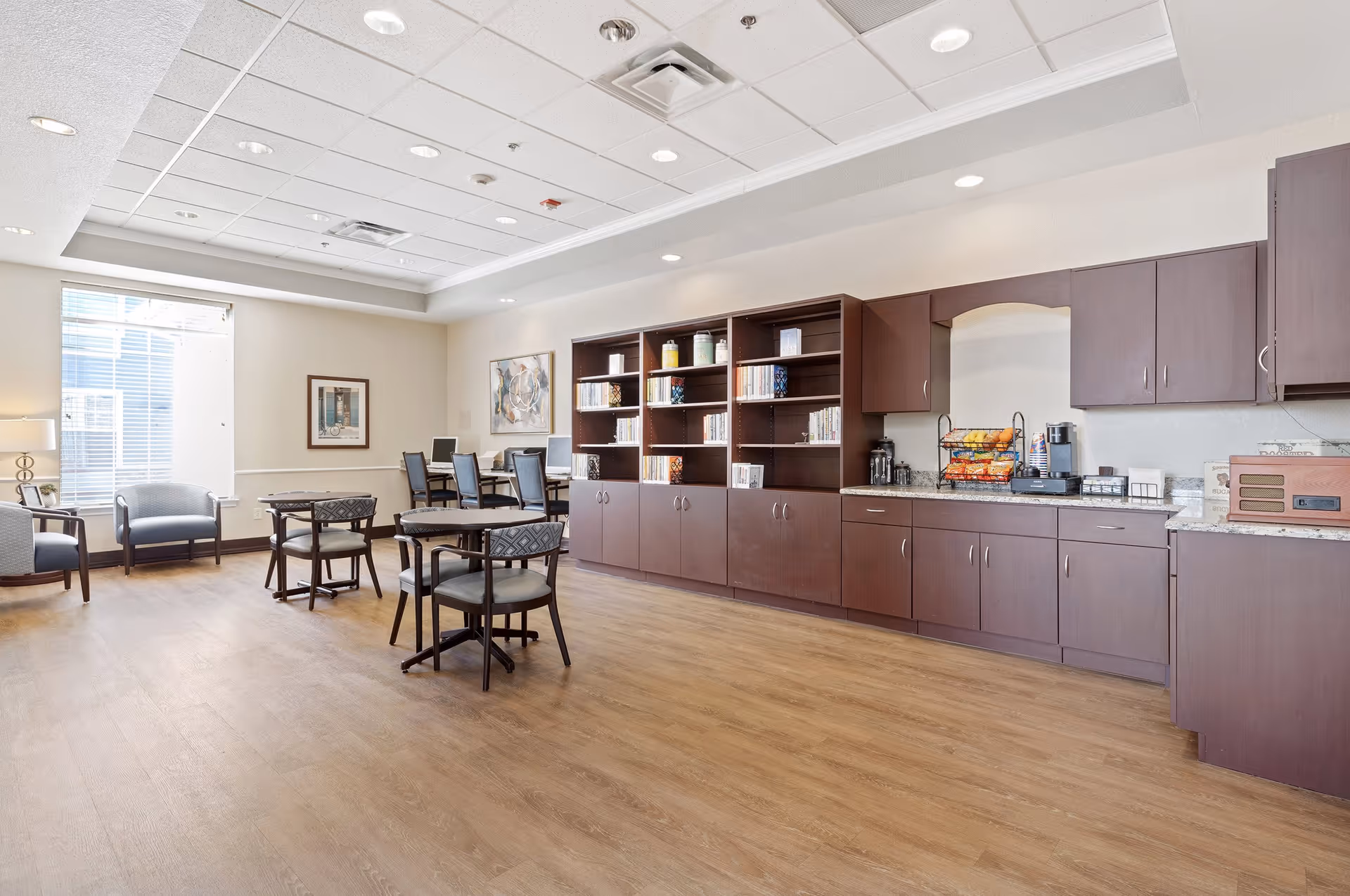 A bright and spacious common area in a senior living facility with wooden flooring, several round tables with chairs, armchairs near a large window, and a long counter with dark wood cabinets and shelves. The counter holds a coffee machine, snacks, and other small appliances. The room has a white ceiling with recessed lighting and framed artwork on the walls.