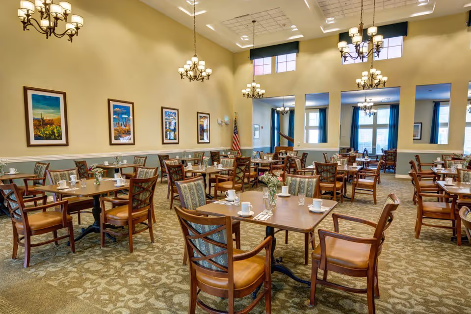A spacious dining room with multiple wooden tables and chairs arranged neatly. Each table is set with white cups, glasses, and silverware. The room features high ceilings with elegant chandeliers, framed artwork on the walls, and large windows with blue curtains allowing natural light to enter. The carpet has a patterned design, and an American flag is visible in the corner.