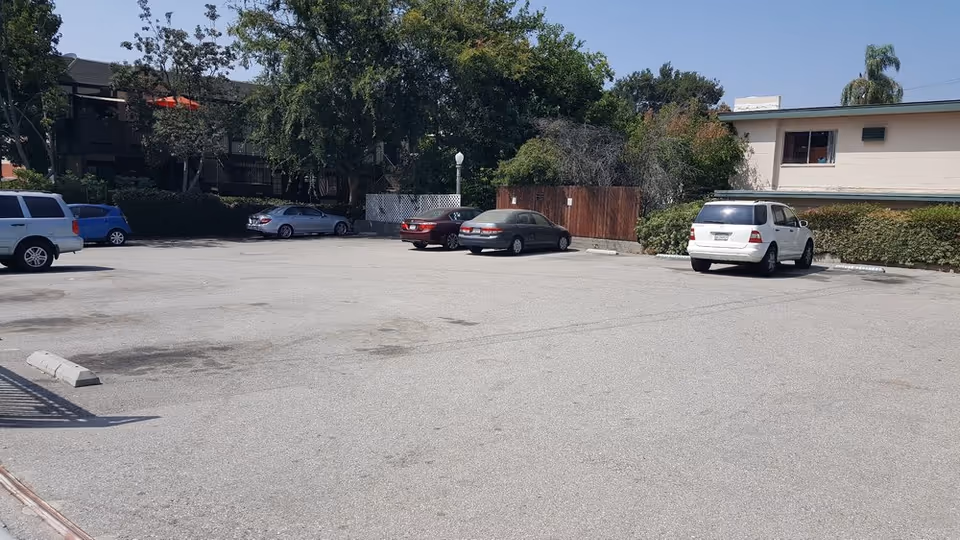 A mostly empty asphalt parking lot with a few parked cars in front of low-rise apartment buildings and trees.
