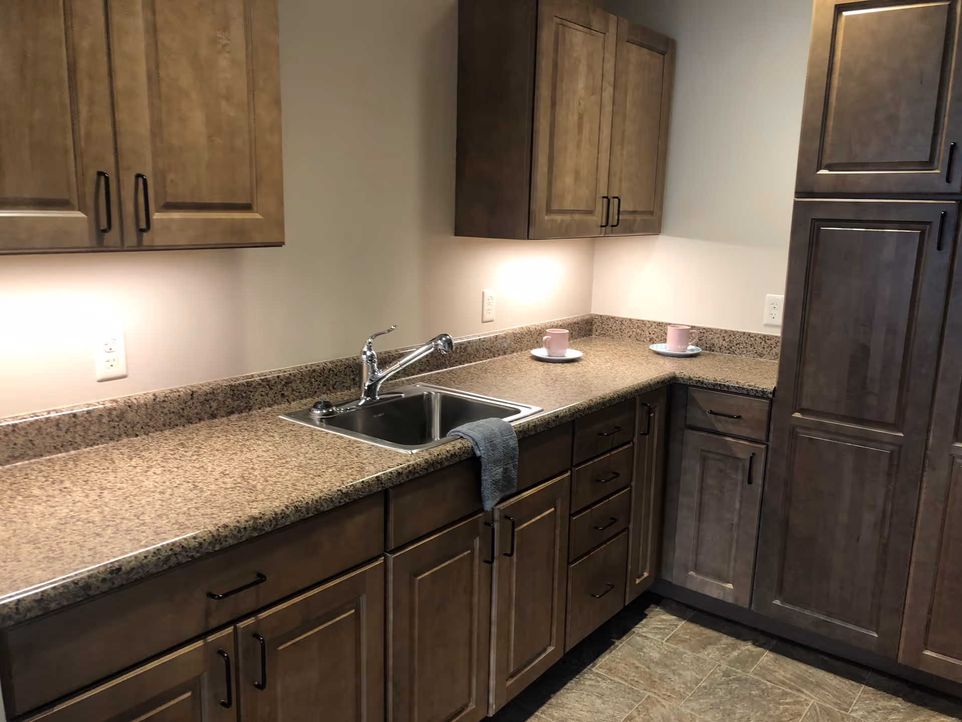 A kitchen area with brown wooden cabinets and drawers, a granite countertop, a stainless steel sink with a faucet, and two pink cups on white saucers placed on the counter. The kitchen has under-cabinet lighting and a tiled floor.