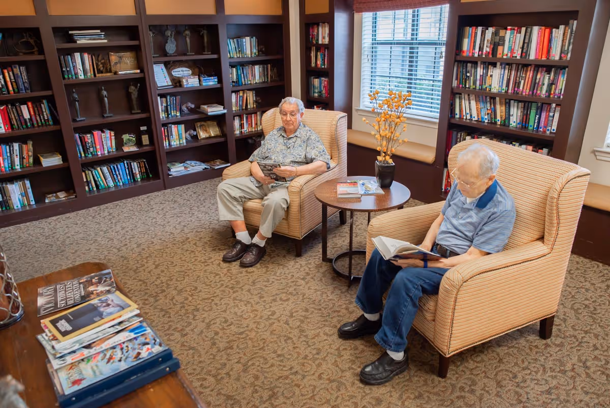 Two elderly men sitting in armchairs in a library or reading room. One man is reading a book while the other is holding a magazine. The room has carpeted floors, bookshelves filled with books and decorative items, a round wooden table with a vase of flowers, and a window with blinds letting in natural light.