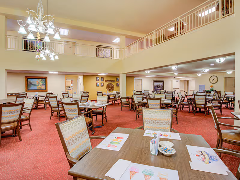 A spacious dining room with multiple tables and chairs arranged neatly. The room has a red carpet, cream-colored walls, and a high ceiling with a chandelier. There are colorful drawings on the tables, a clock on the far wall, and framed pictures decorating the walls.