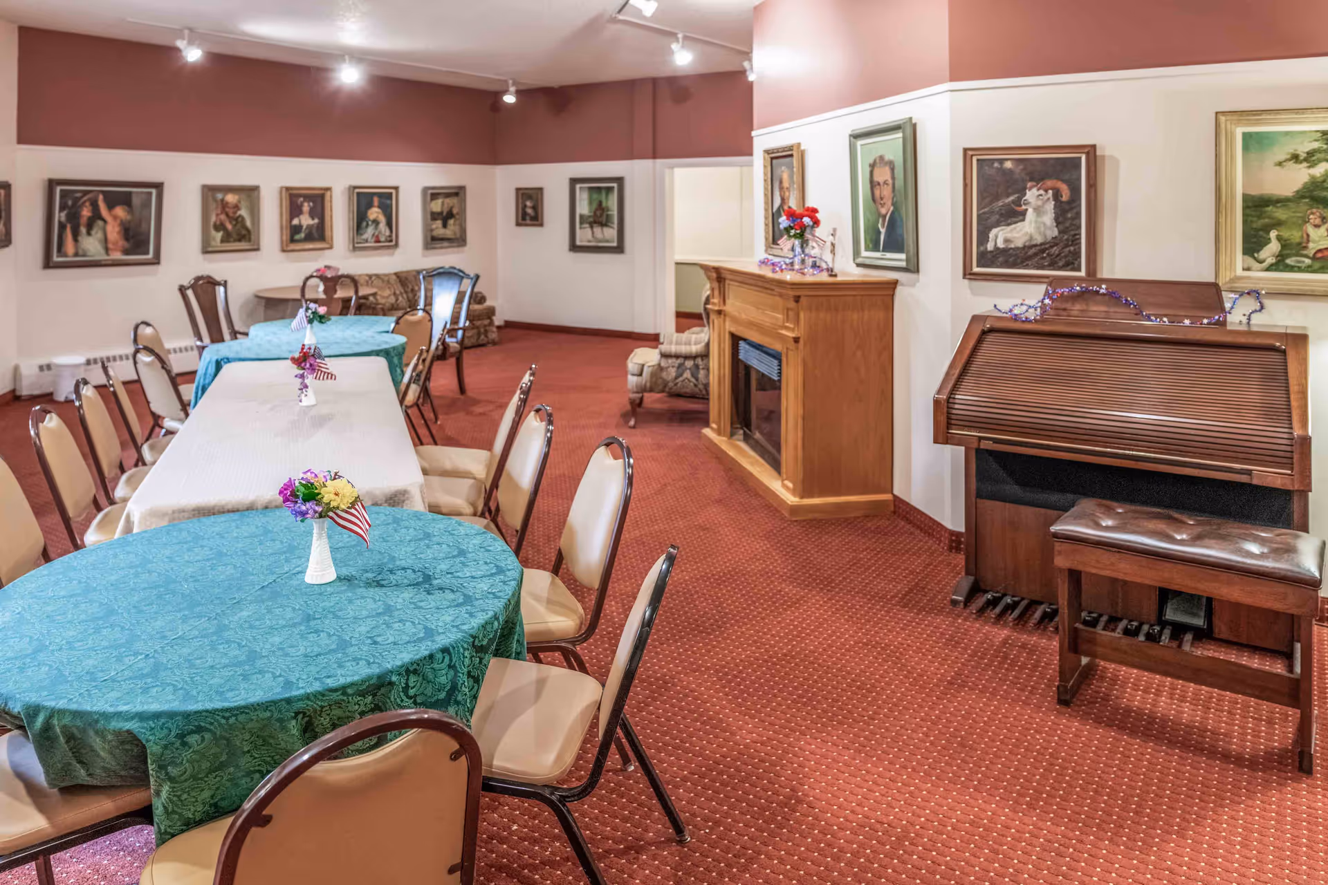 Dining/activity room with round and rectangular tables set with centerpieces, chairs, a piano/organ, a fireplace, and framed paintings on the walls.