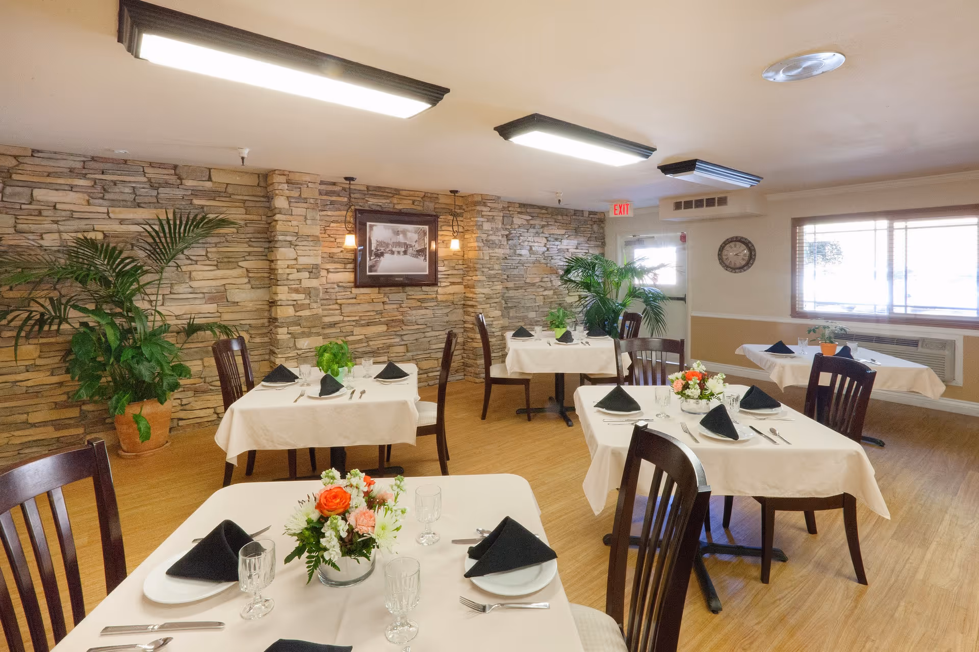 A dining room with several tables covered in white tablecloths, each set with plates, silverware, glasses, and black folded napkins. The room features a stone accent wall with a framed picture, potted plants, wooden chairs, and large windows letting in natural light. The floor is wooden, and ceiling lights illuminate the space.