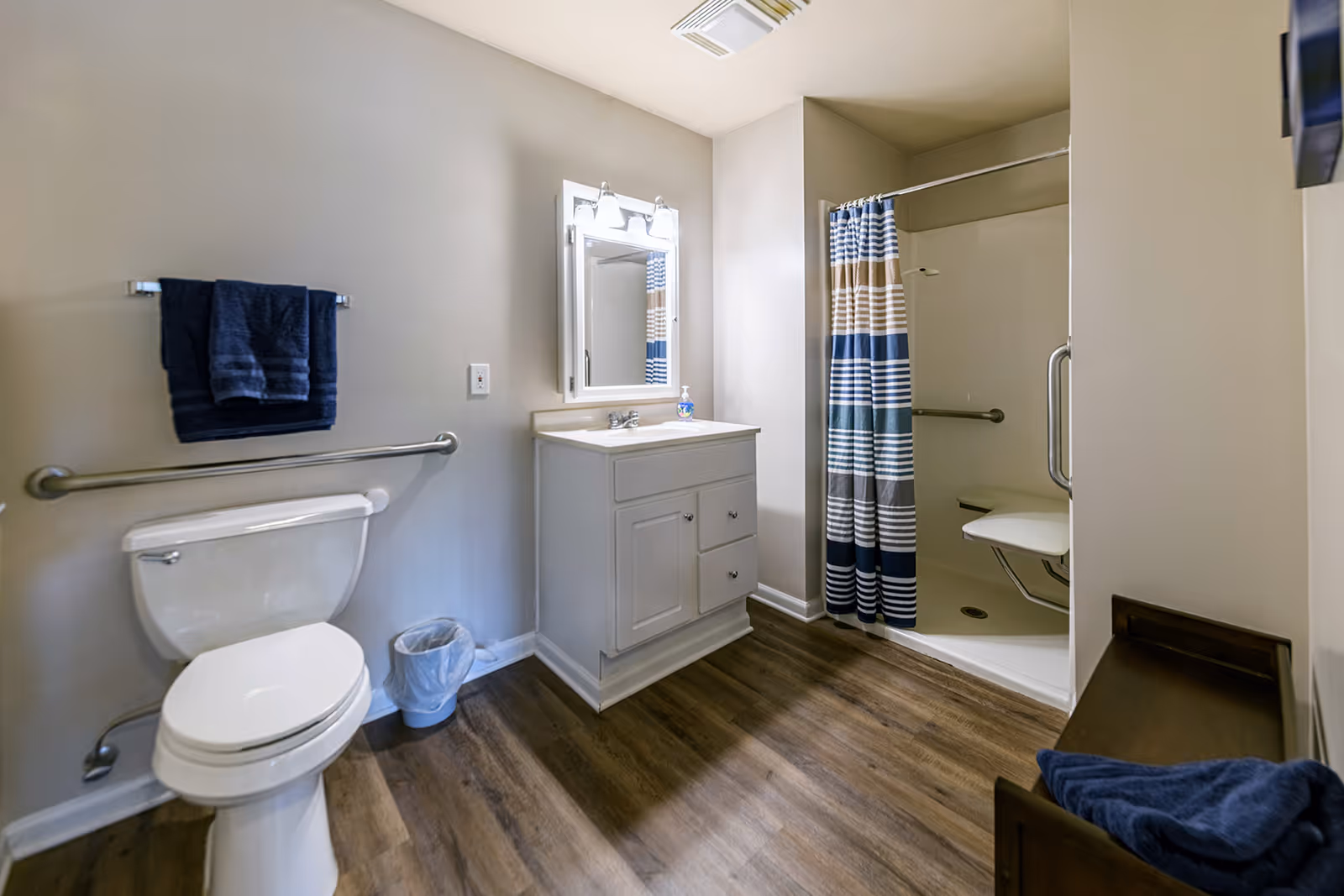 A clean and accessible bathroom with a white toilet, a vanity with a sink and mirror above it, a shower with a striped blue and beige curtain, and grab bars for safety. The floor is wood-style, and there are blue towels hanging and folded on a wooden bench.