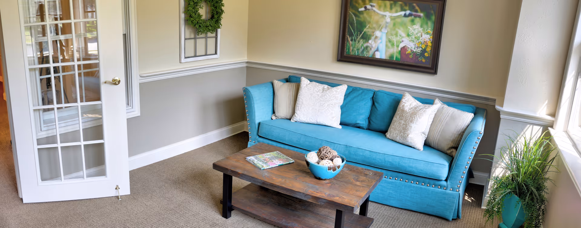 A cozy living room area featuring a turquoise sofa with white and beige pillows, a wooden coffee table with decorative items and a magazine, a framed picture of birds on the wall, a green wreath hanging above a window, and a potted plant near a window letting in natural light.