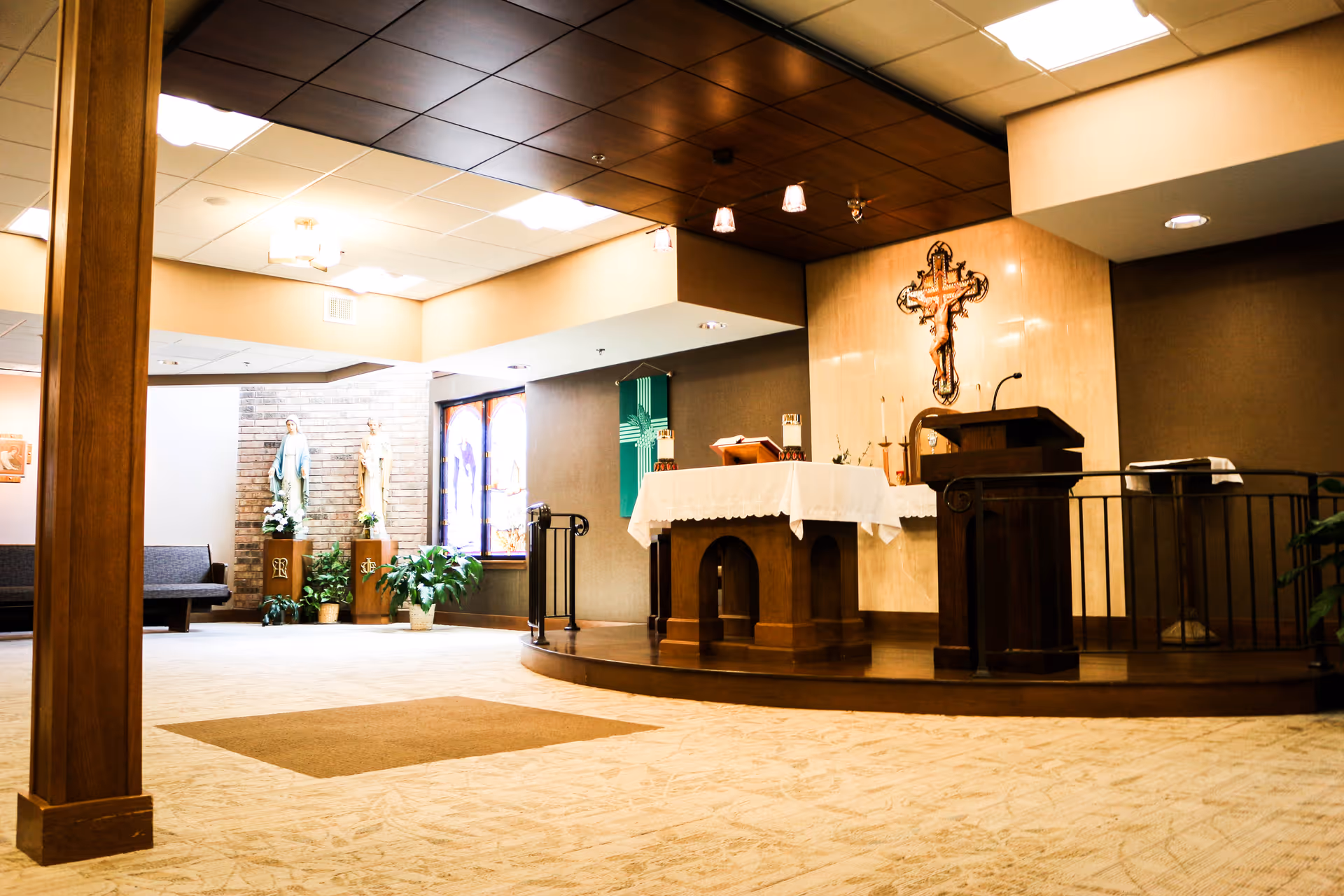 Interior view of a chapel area in a senior living facility with an altar covered by a white cloth, a wooden lectern, a crucifix mounted on the wall, statues of religious figures, stained glass windows, and potted plants.