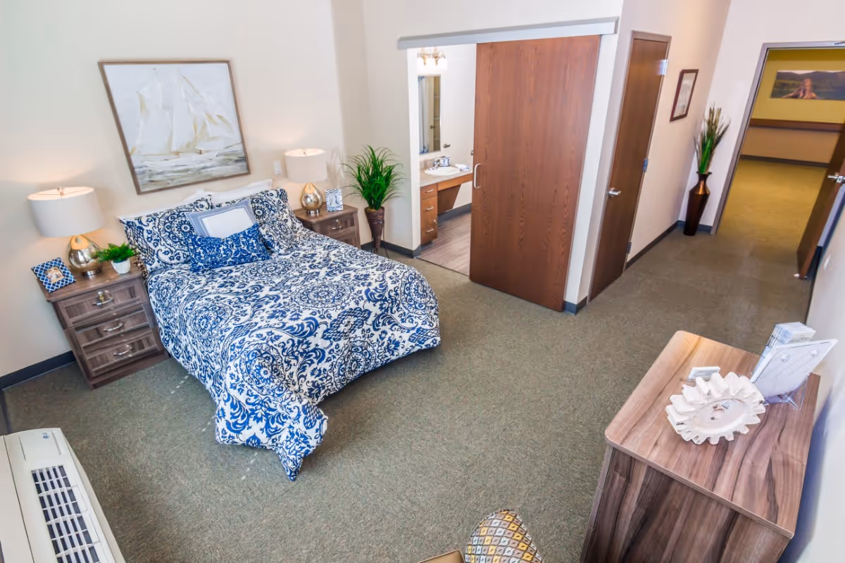 A neatly arranged bedroom with a bed covered in blue and white patterned bedding, two bedside tables with lamps and small plants, a large painting of a sailboat on the wall, and a sliding wooden door leading to a bathroom with a sink and mirror. The room has carpeted flooring and a hallway visible in the background with a plant in a tall vase.