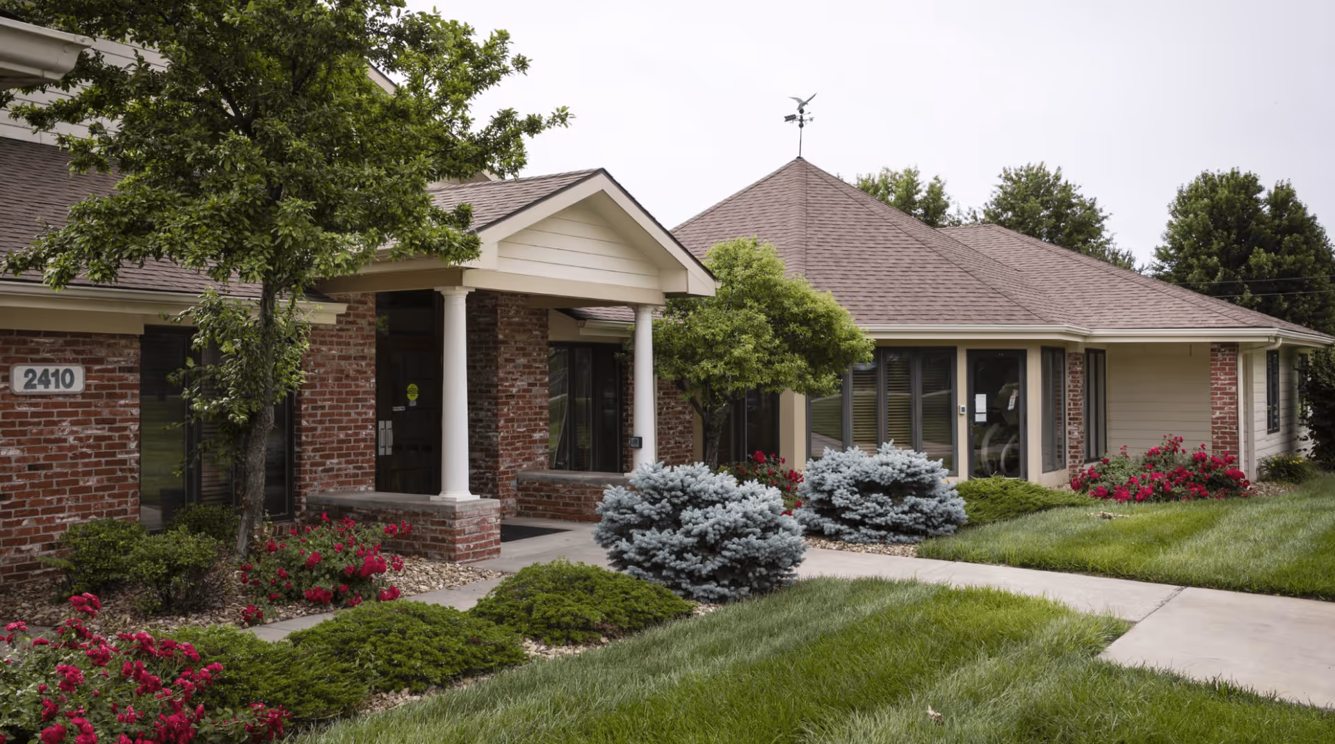 Front entrance of a single-story brick assisted living building with columns, landscaped shrubs, and the address number 2410.