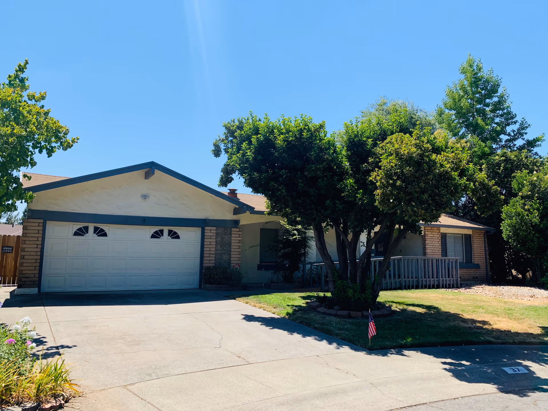 Front exterior view of a single-story residential building with a two-car garage, a driveway, and a well-maintained lawn with trees and shrubs under a clear blue sky.