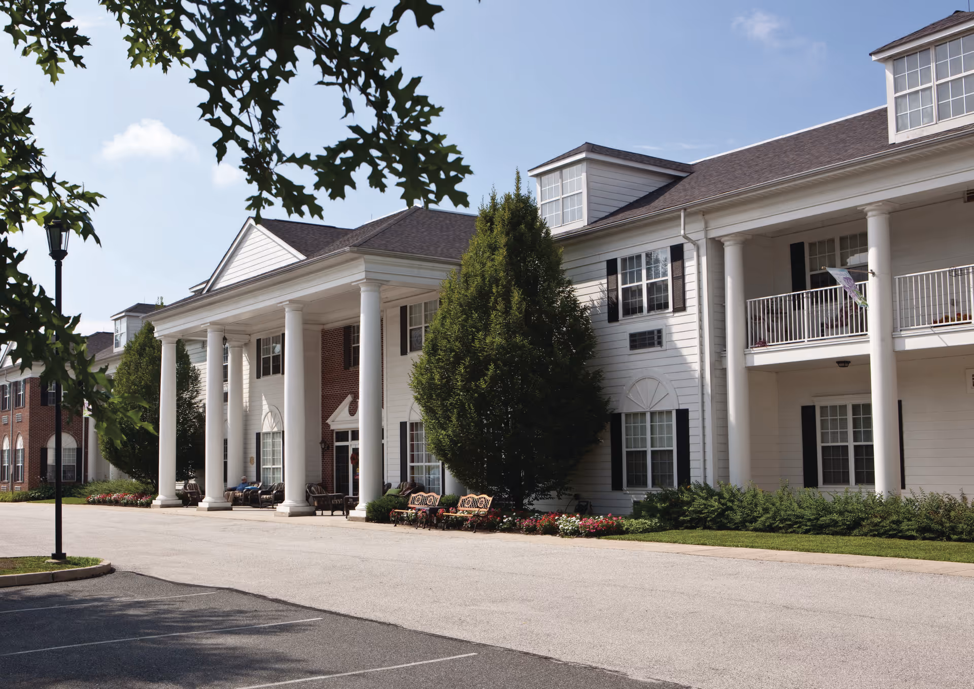 Exterior view of Blue Bell Place, a senior living facility with white columns, balconies, and a well-maintained garden area in front. The building has a classic architectural style with large windows and a paved driveway.