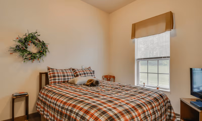 A cozy bedroom with a bed covered in a red, white, and black plaid comforter and matching pillows. A small stuffed animal rests on the bed. There is a window with blinds and a beige valance letting in natural light. A decorative green wreath with small red berries hangs on the wall above a small wooden side table with a few items on it. A small wooden chair is placed in the corner near the window, and a TV is on a stand to the right of the bed.