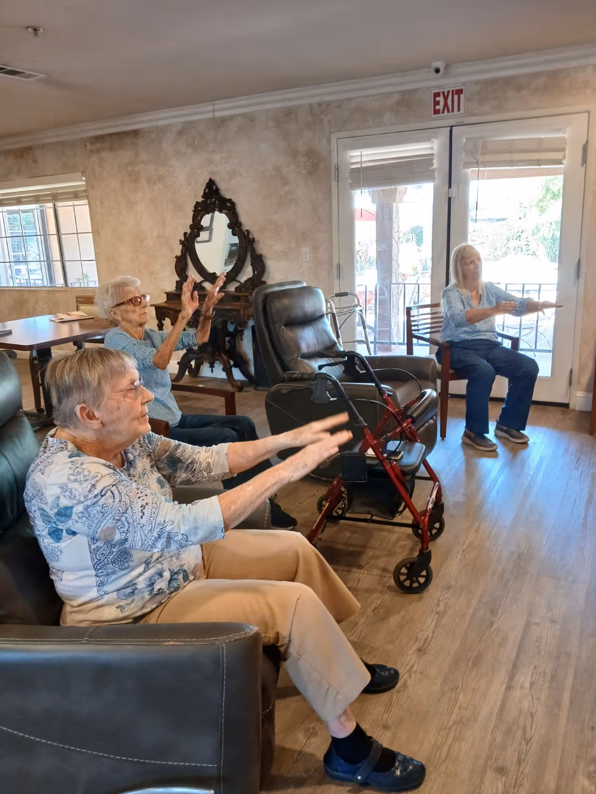 Three elderly women seated in a living room area of an assisted living facility, engaging in a seated exercise with their arms extended forward. The room has wooden flooring, a large window, a decorative mirror on the wall, and a glass door with an exit sign above it. One woman uses a red walker placed beside her chair.
