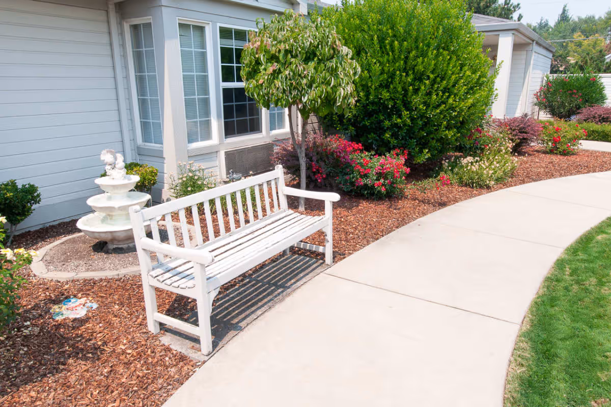 A white wooden bench next to a concrete pathway in a garden area outside a building. The garden has mulch, various green bushes, flowering plants, and a small white tiered water fountain near the building's window.