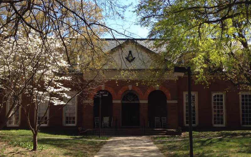 Front exterior view of a brick building with arched doorways and multiple windows, partially shaded by trees with green and white blossoms, with a paved walkway leading to the entrance.