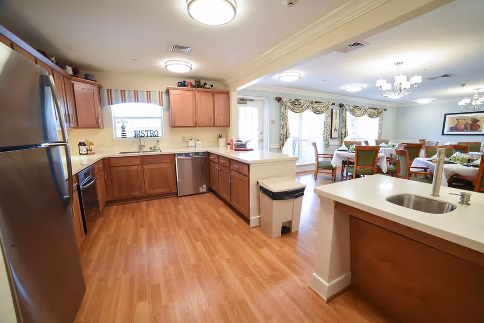 A spacious kitchen area with wooden cabinets, stainless steel refrigerator, oven, and dishwasher. The kitchen has a light wood floor and white countertops. Beyond the kitchen is a dining area with tables covered in white tablecloths and green chairs, with large windows letting in natural light and floral curtains.