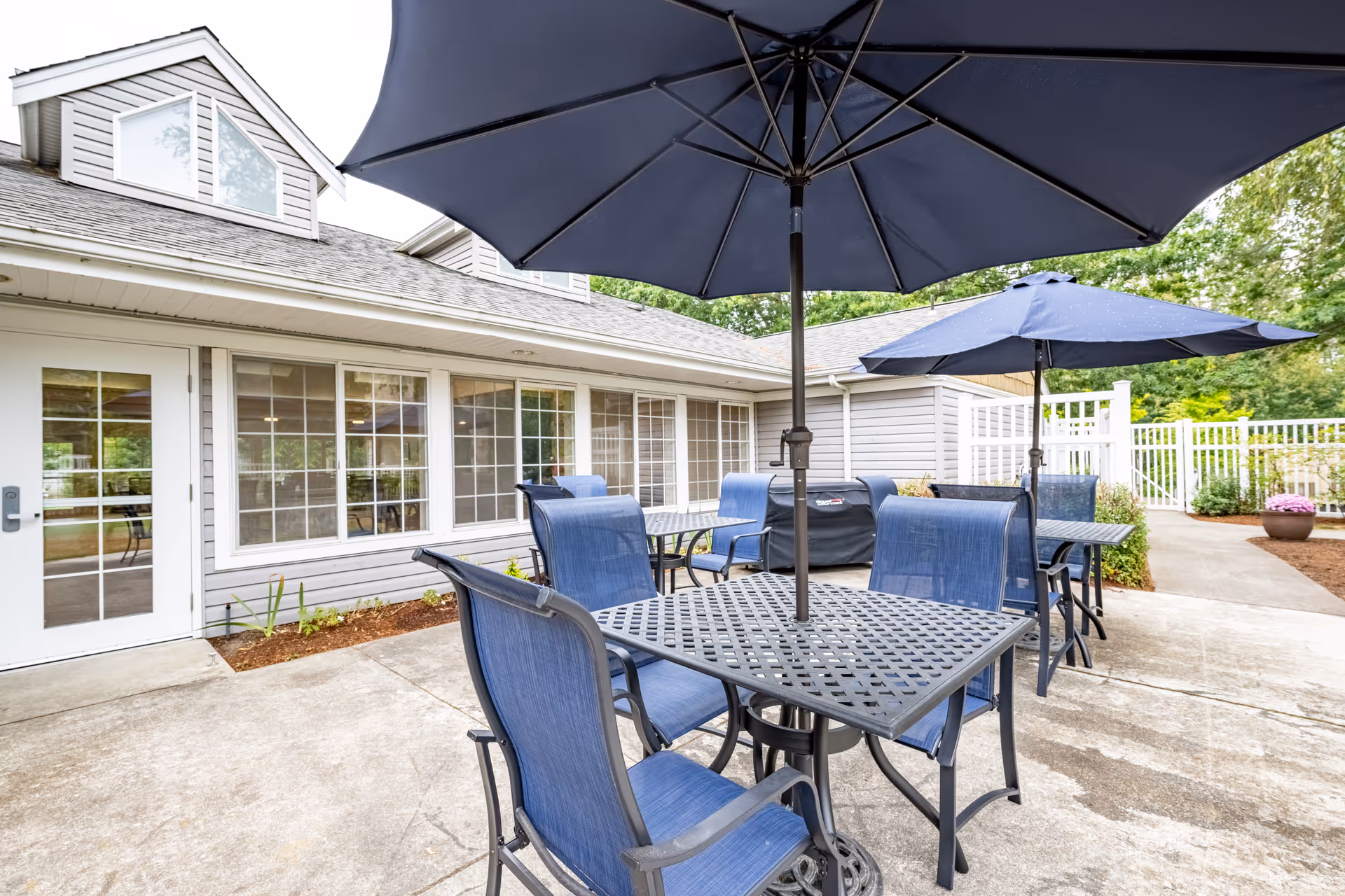 Outdoor patio area at Birchview Memory Care with metal tables and blue chairs under large blue umbrellas, adjacent to a building with multiple windows and a white door, surrounded by greenery and a white fence.