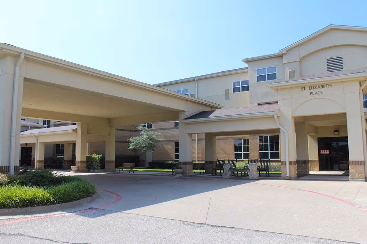 Exterior view of the entrance to a senior living facility named St. Elizabeth Place, showing a covered drop-off area with benches and landscaping under a clear blue sky.