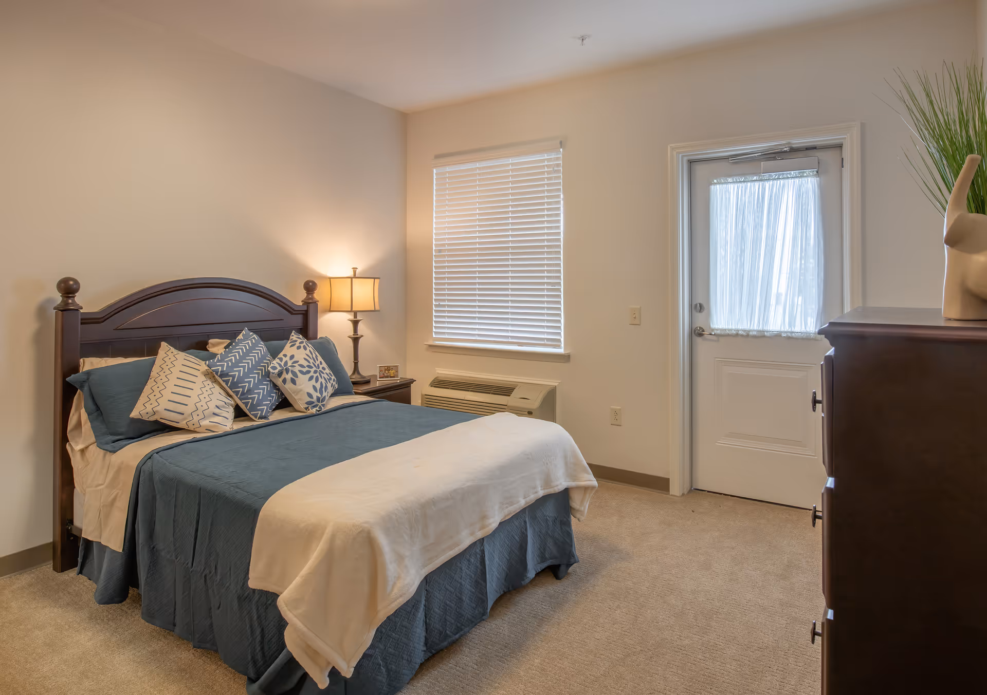 A tidy bedroom with a wooden bed dressed in blue and white bedding, a nightstand with a lamp, a window with blinds, and a door.