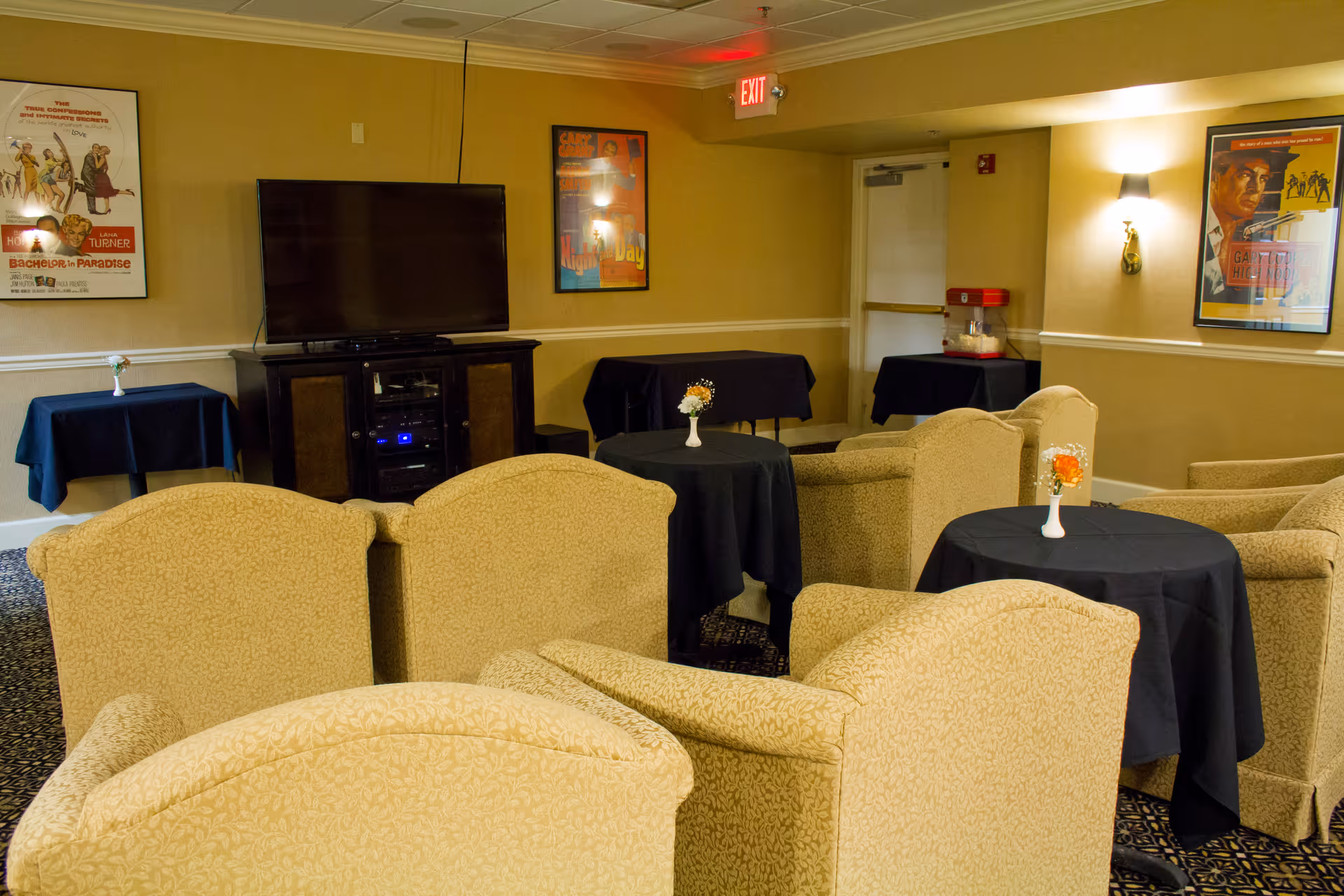 A cozy lounge area with beige upholstered armchairs arranged around small round tables covered with black tablecloths and decorated with small flower vases. A large flat-screen TV is mounted on a dark wooden cabinet against the wall. The walls are adorned with framed vintage movie posters, and there is a popcorn machine on a table in the corner. The room has warm lighting and an exit sign above a door.