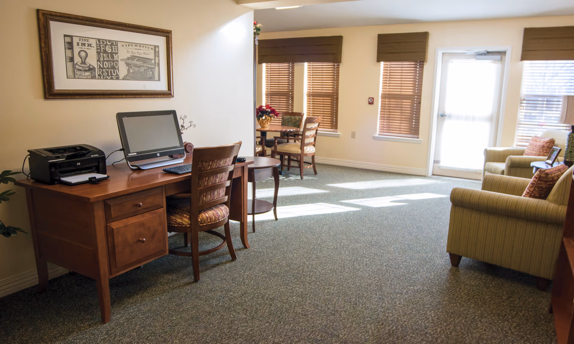 Sunlit common room with a wooden desk and computer, a small dining table, and upholstered armchairs near windows and a glass door.