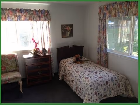 A small bedroom with a single bed covered in a floral bedspread. There is a teddy bear and a doll placed on the bed. Next to the bed is a wooden nightstand with three drawers, a small globe, and a flower arrangement on top. The room has two windows with floral curtains, and a floral upholstered chair is positioned near one window. A framed picture hangs on the wall above the bed.