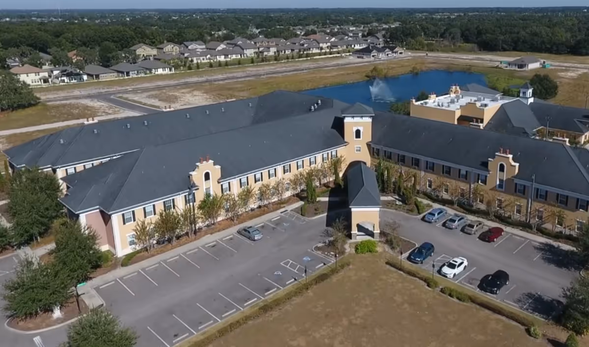 Aerial view of a two-story assisted living building with a central porte-cochere, surrounding parking lot, and a pond with a fountain nearby.