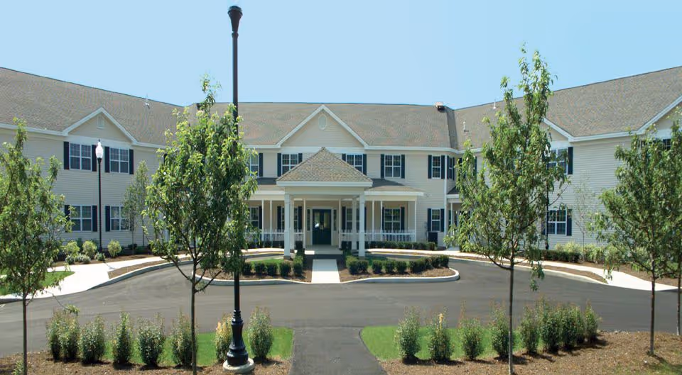 Front exterior of a two-story senior living facility with a covered entrance, circular driveway, and landscaped trees and shrubs.