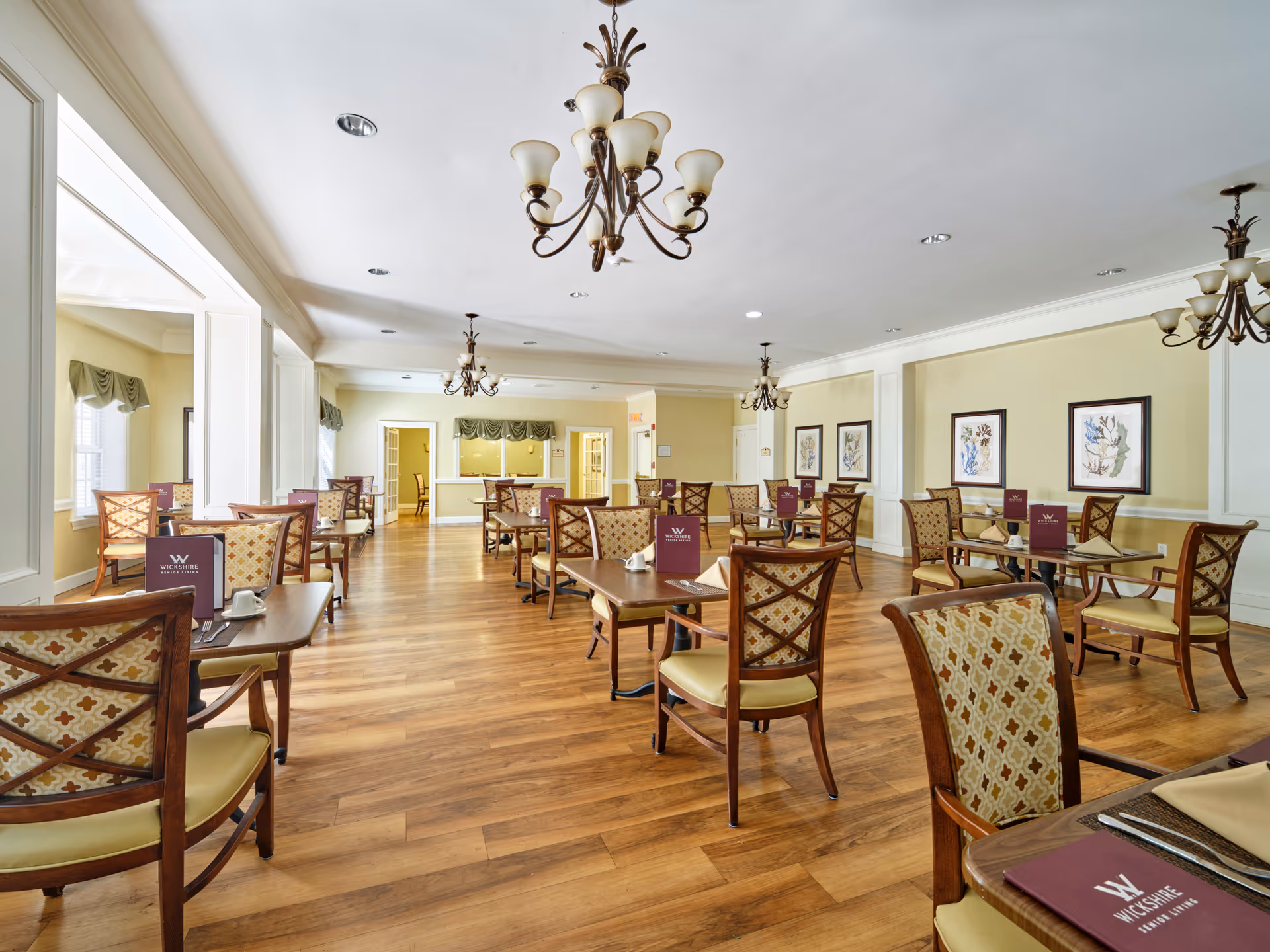 A spacious dining room with wooden floors, multiple tables and chairs arranged neatly, chandeliers hanging from the ceiling, and framed artwork on the walls. Each table has a maroon menu with the text 'Wickshire Senior Living'.