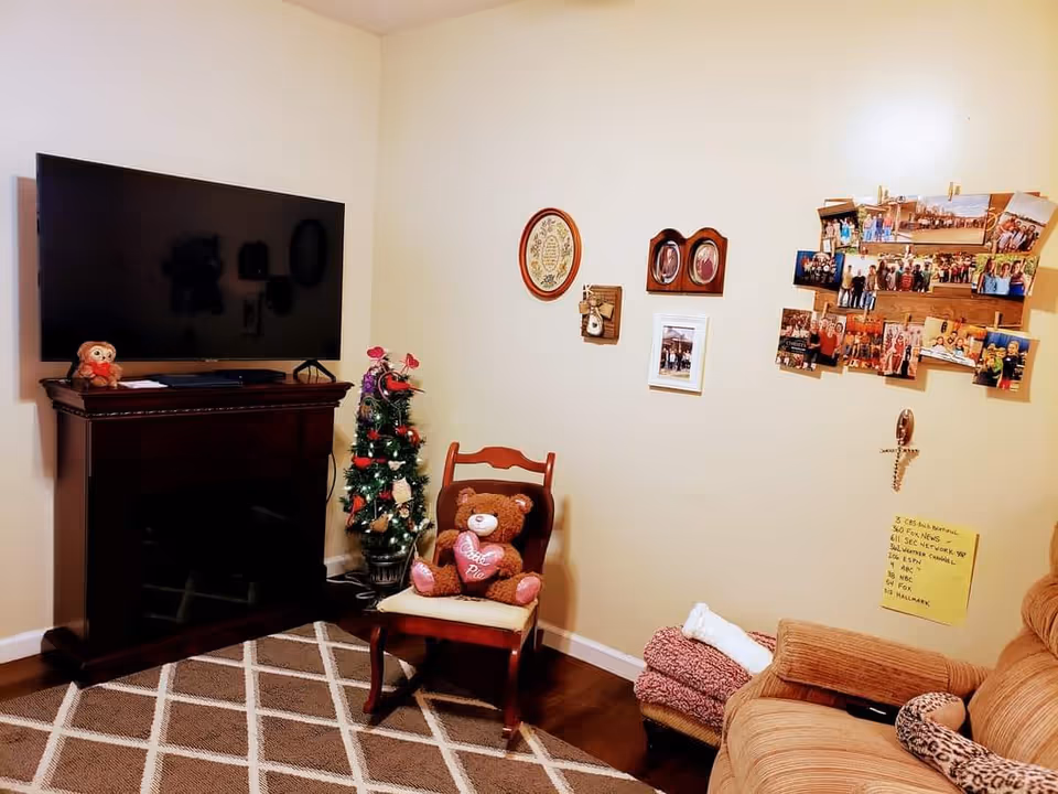 A cozy living room corner with a large flat-screen TV mounted on a dark wooden stand, a small decorated Christmas tree, a wooden chair with a teddy bear holding a heart-shaped pillow, a beige armchair with folded blankets and a neck pillow, and a wall decorated with framed pictures and a collage of photographs.