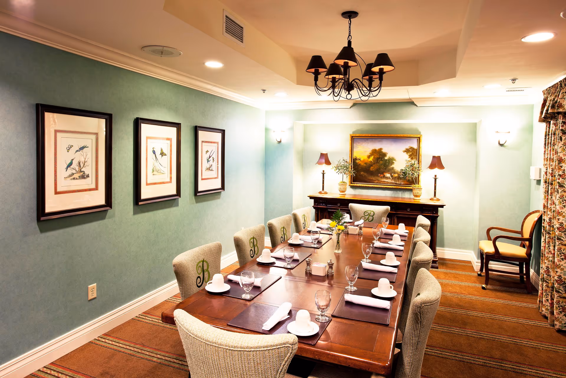 A formal dining room with a long wooden table set for ten people. Each place setting includes a white cup and saucer, a glass, and a rolled white napkin on a dark placemat. The chairs around the table are upholstered in a light fabric with a green 'B' embroidered on the backrests. The walls are painted light green and decorated with framed bird illustrations on one side and a landscape painting above a wooden sideboard on the other. Two table lamps with dark shades and small potted plants are on the sideboard. A black chandelier hangs from the ceiling, and floral curtains cover a window on the right side of the room.