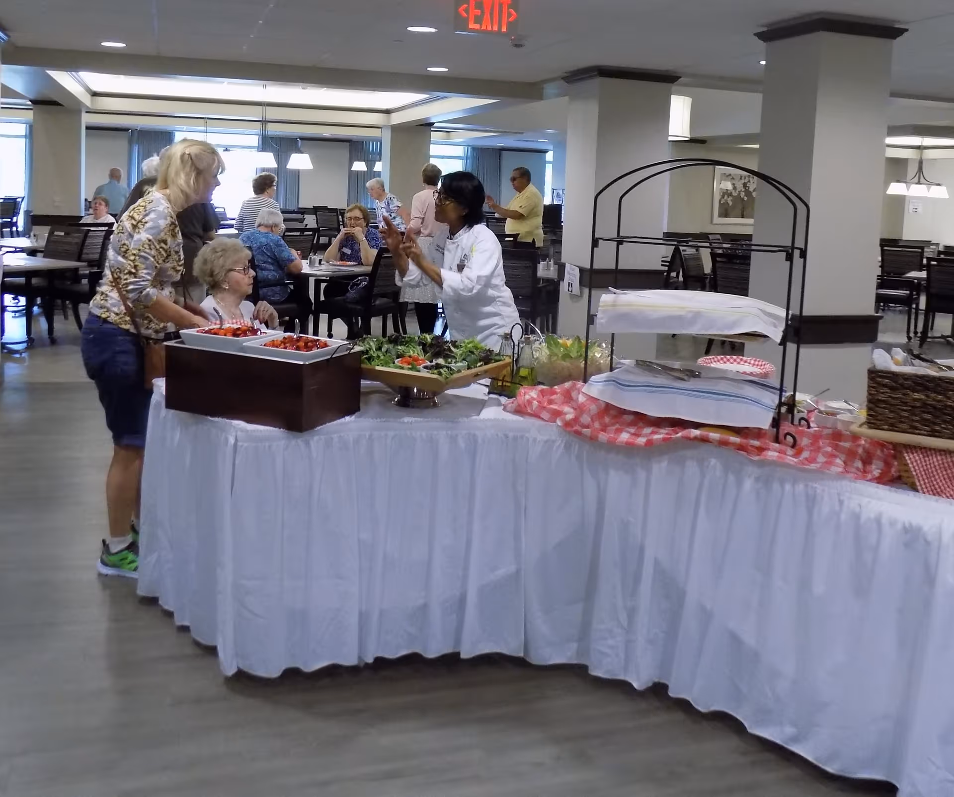 A dining area in a senior living facility with several elderly people seated at tables and a buffet table in the foreground covered with a white tablecloth and various food items. A staff member in a white uniform is interacting with two women near the buffet. The room has modern lighting and a clean, spacious layout.
