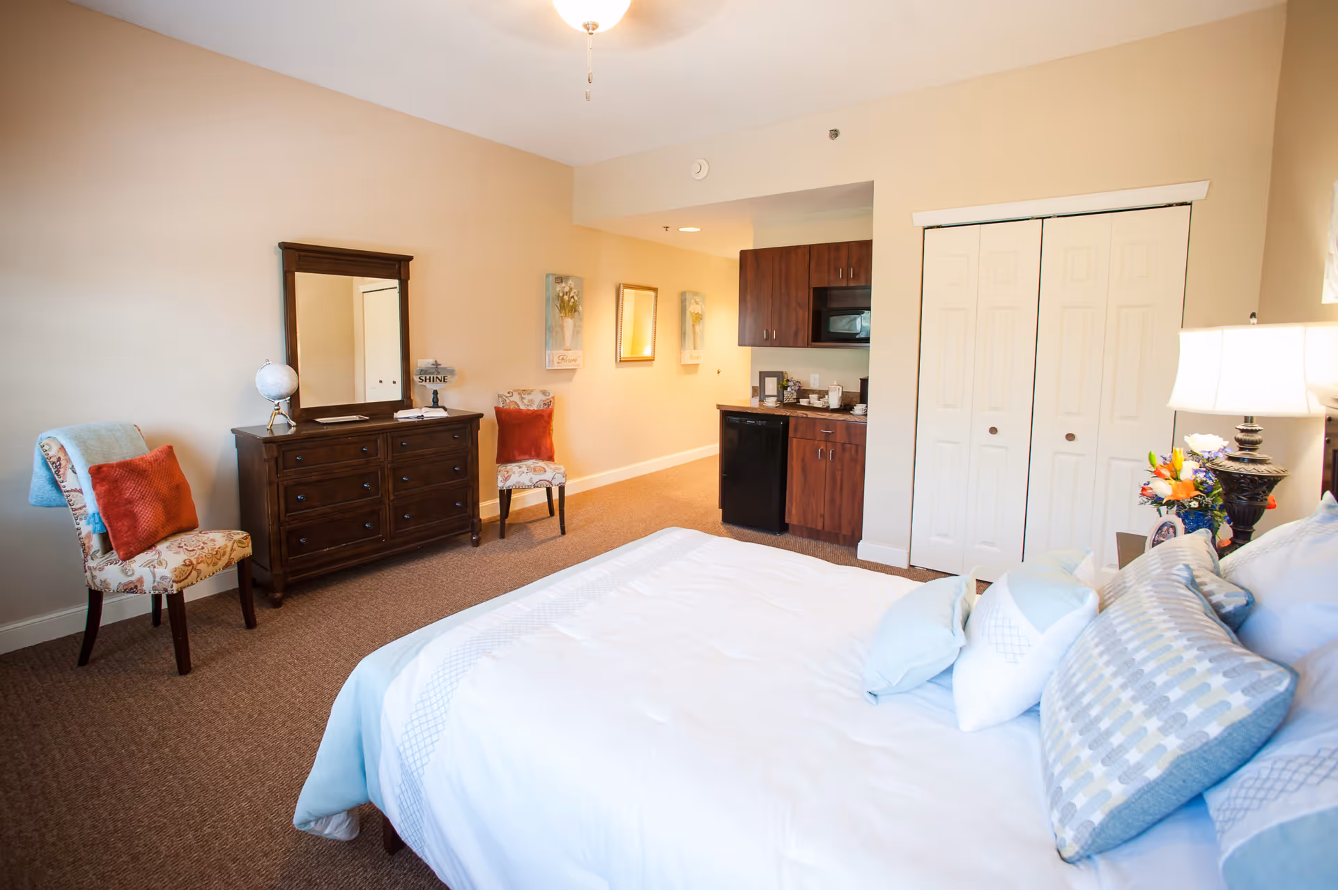 Bright furnished senior living bedroom with a bed, dresser and chairs, plus a small kitchenette and closet.