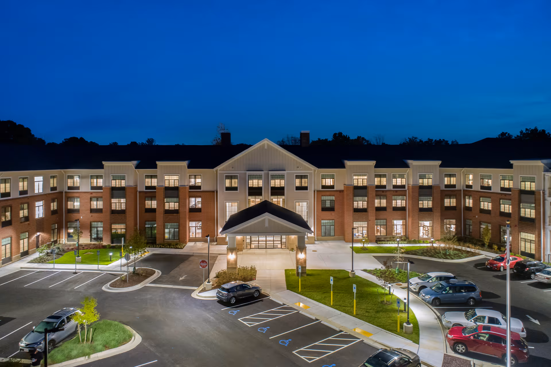 Nighttime exterior view of a large, well-lit senior living facility building with multiple windows and a covered entrance. Several cars are parked in the parking lot, including designated handicapped parking spaces. The sky is dark blue, indicating evening or early night.