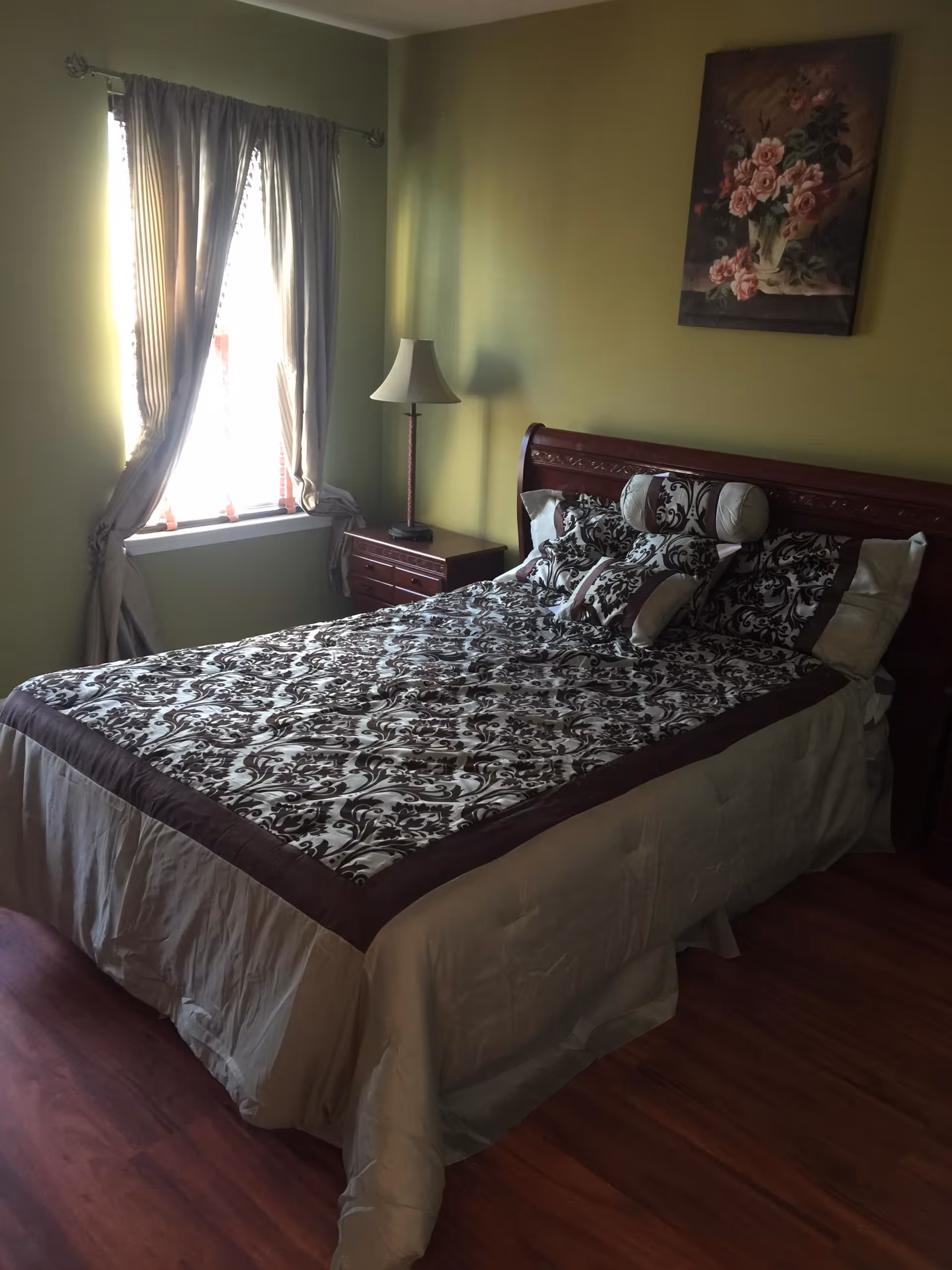 A bedroom with a wooden bed frame and a bed covered with a patterned bedspread and multiple decorative pillows. There is a wooden nightstand with a lamp next to the bed. A window with curtains is on the left wall, and a floral painting hangs above the bed on the green wall.