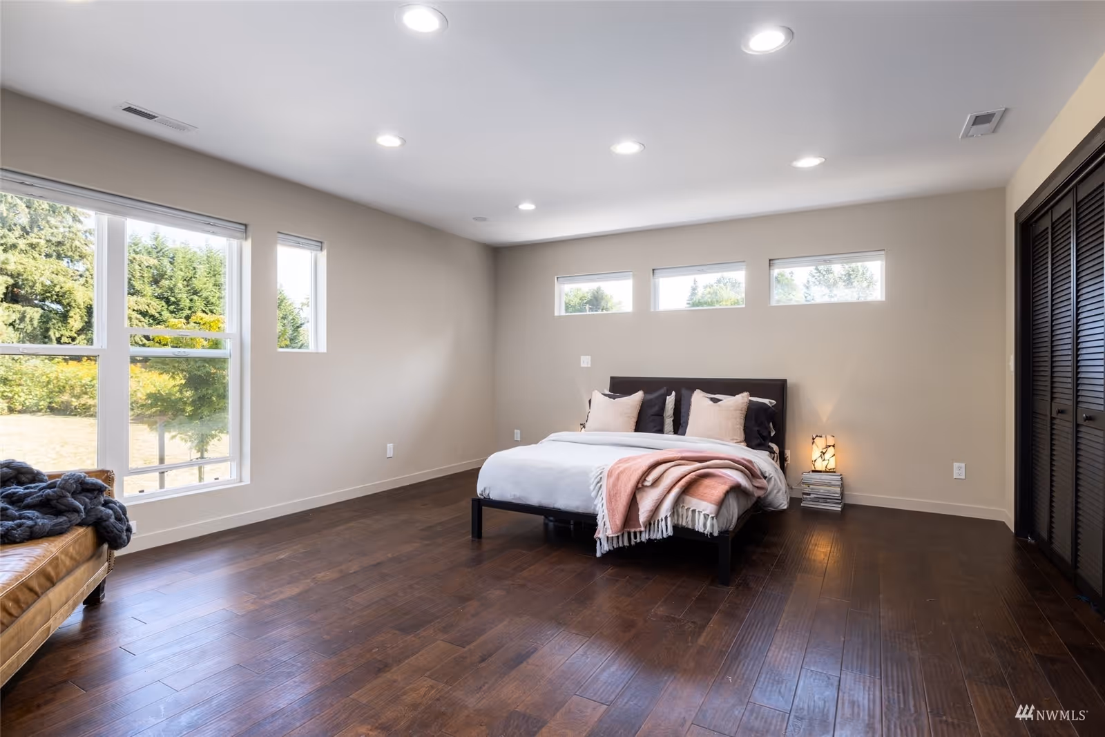 Spacious minimalist bedroom with a bed, hardwood floors, multiple windows, and neutral-colored walls.