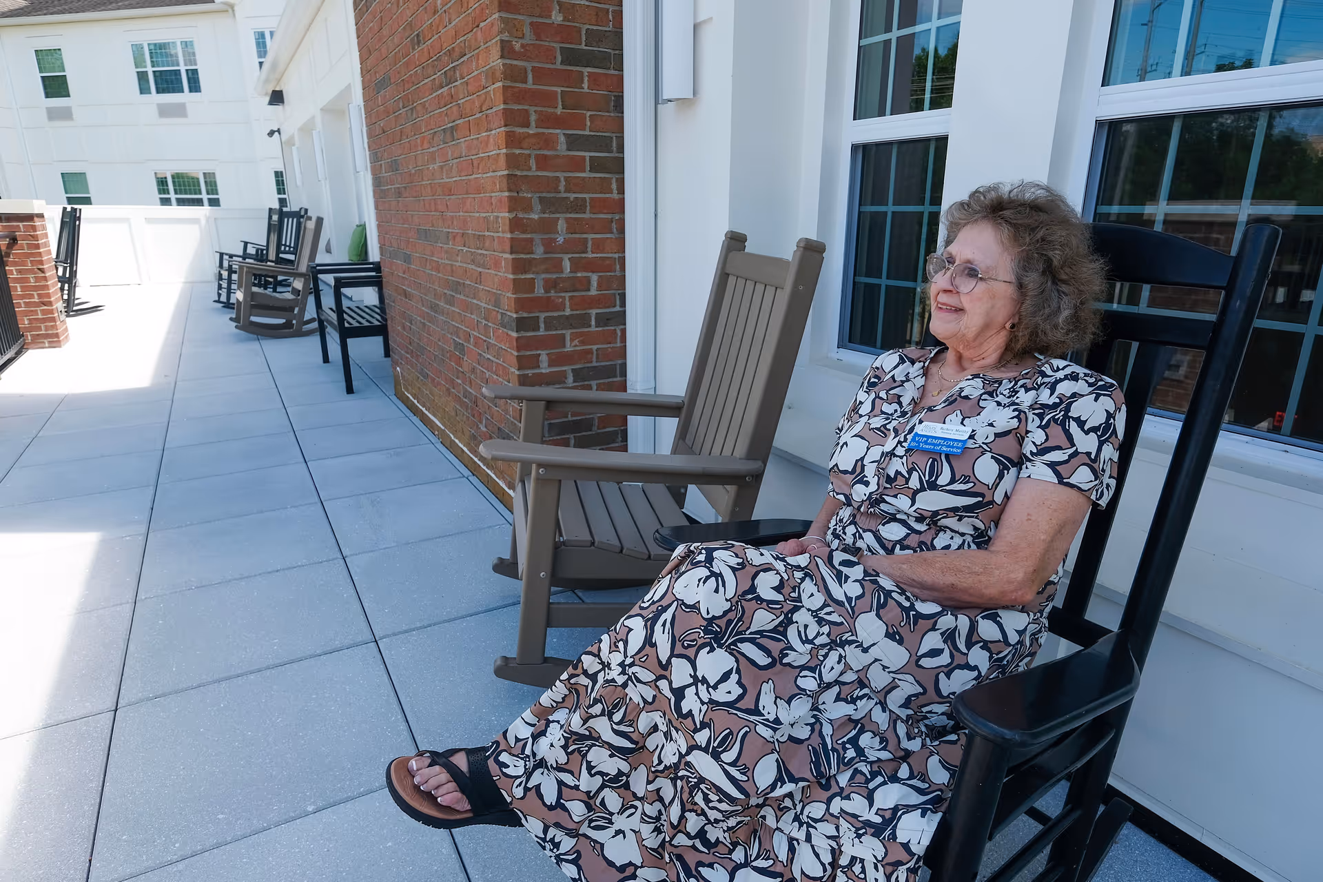 A smiling elderly woman in a floral dress sits on a rocking chair on an outdoor patio beside a brick wall and windows.