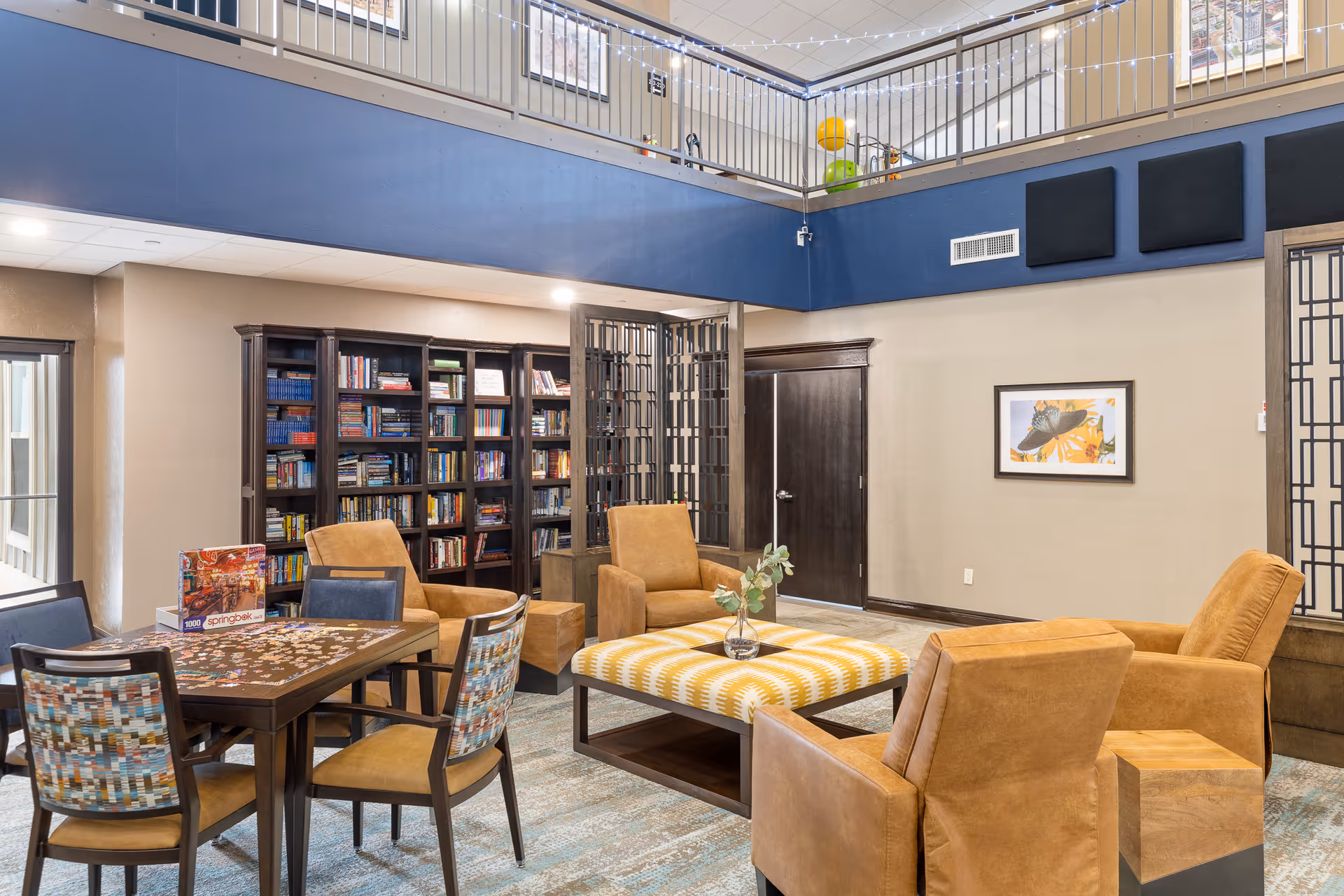 A cozy common area in a senior living facility with a high ceiling and an upper balcony. The room features a puzzle table with four chairs, a large ottoman with a vase on top, and four comfortable armchairs arranged around it. There is a bookshelf filled with books against the wall, a framed picture of a bird on the wall, and decorative panels on the upper part of the wall.