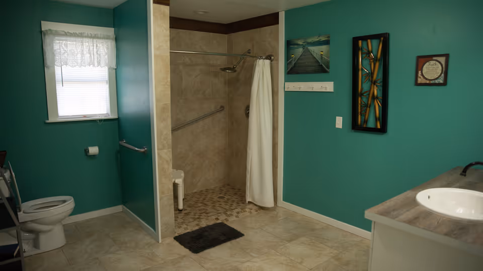 A bathroom with teal walls featuring a toilet near a window with a lace curtain, a walk-in tiled shower with a white curtain and grab bars, a dark bath mat on the floor, and a sink with a wooden countertop. The walls are decorated with framed pictures and a coat rack.
