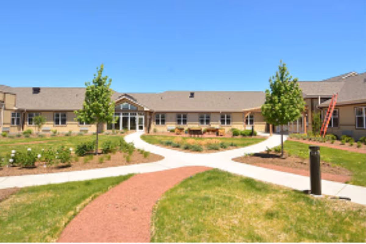 Outdoor view of Silverado Frederick Memory Care Community showing a single-story building with a beige exterior and a brown roof. There are multiple windows and a central entrance with glass doors. The foreground features a landscaped garden with green grass, small trees, shrubs, and intersecting paved walkways under a clear blue sky.