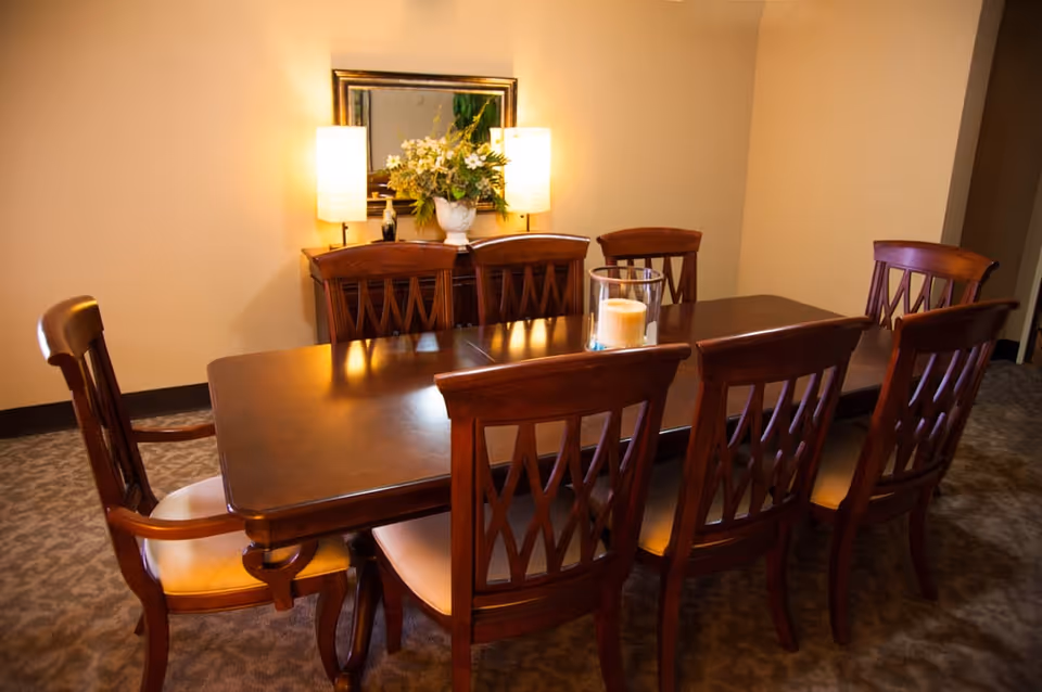 A dining room with a polished wooden table surrounded by eight matching wooden chairs with cushioned seats. On the table is a glass candle holder with a candle inside. Against the wall is a sideboard with two lit lamps and a floral arrangement in a white vase, with a framed mirror hanging above it. The room has beige walls and carpeted flooring.