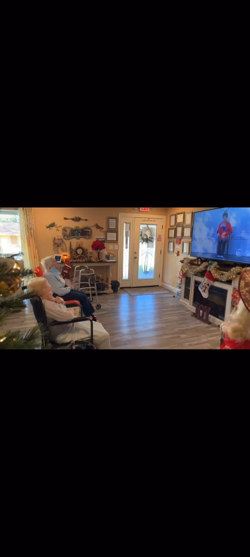 Elderly residents in wheelchairs sit in a decorated common room watching a television above a fireplace.